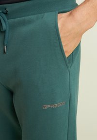 Mano descansando dentro del bolsillo delantero de unos pantalones de chándal de la marca Freddy, color teal, con la cinturilla ajustable de cordón visible.