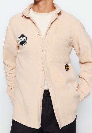 Persona che indossa una camicia in pile beige con bottoni, taschino sul petto e toppe con le scritte "DREAM WORLD" e "BEYOND".