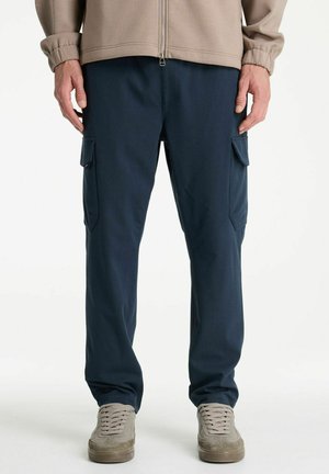Man met marineblauwe cargobroek met zijvouwzakken, beige jas met rits en beige suède sneakers, staand op een witte achtergrond.