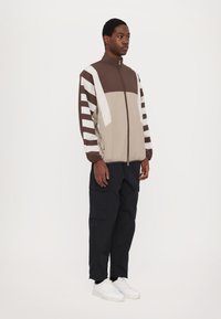 Hnědo-béžová bunda na zip s vertikálními pruhy, doplněná černými cargo kalhotami a bílými teniskami. Hladká látka, design s vysokým límcem.