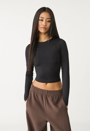 Schwarzes, langärmliges, cropped Top mit rundem Ausschnitt, kombiniert mit braunen Hosen mit elastischem Bund, die eine glatte Textur und eine figurbetonte Passform aufweisen.