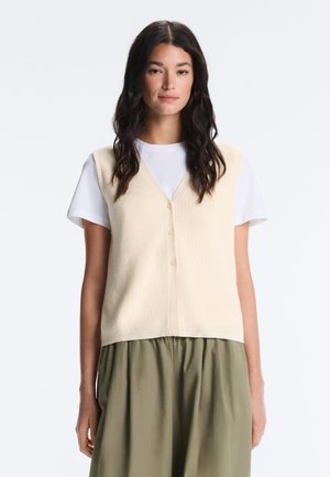 Femme aux longs cheveux foncés portant un gilet crème boutonné sur une chemise blanche à manches courtes et une jupe taille haute vert olive, debout sur fond blanc.