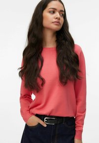 Pull corail à manches longues avec un col rond et des poignets côtelés, associé à un pantalon en denim foncé et une ceinture noire avec une boucle argentée.