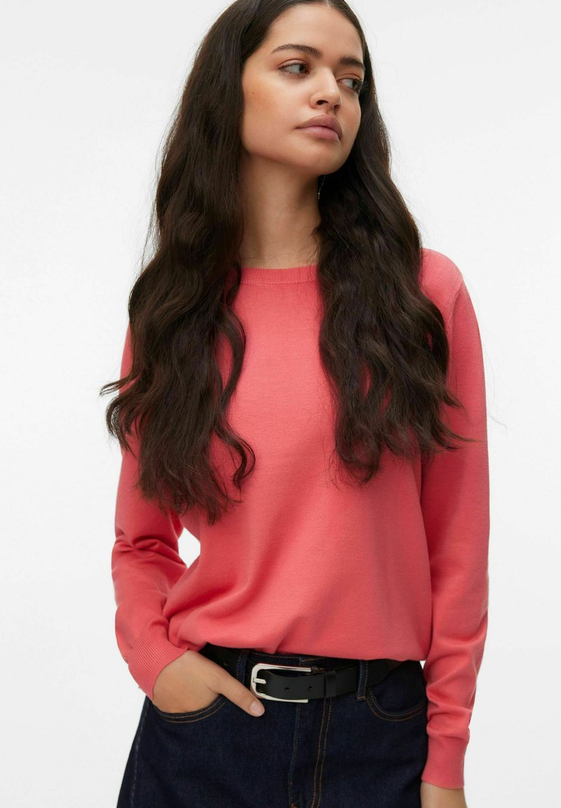 Pull corail à manches longues avec un col rond et des poignets côtelés, associé à un pantalon en denim foncé et une ceinture noire avec une boucle argentée.