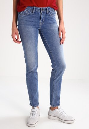 Jean en denim bleu clair avec une coupe slim, comportant cinq poches et un léger délavage. Porté avec des baskets blanches et une chemise rouge.