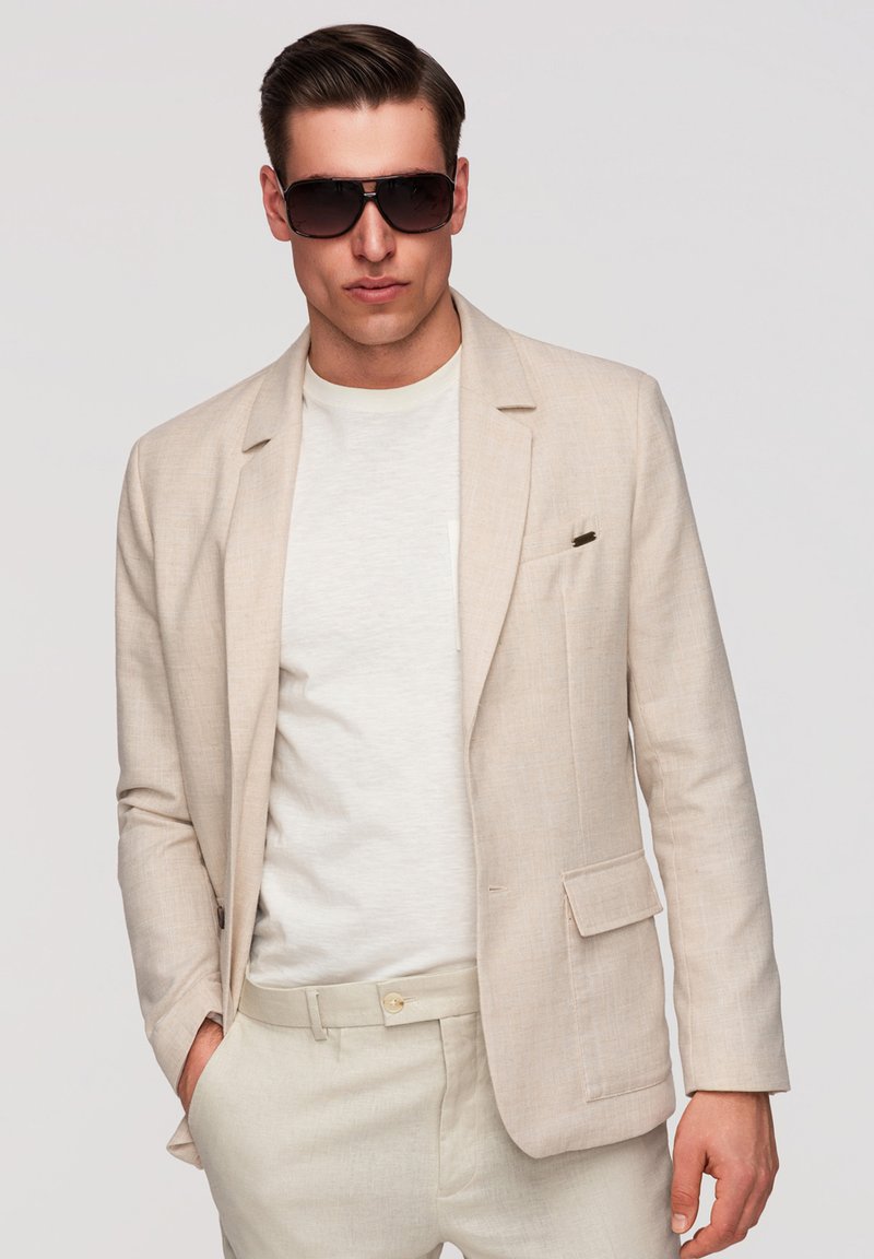 Homme portant un costume beige, une chemise blanche et de grandes lunettes de soleil foncées, debout avec une main dans la poche sur un fond uni.