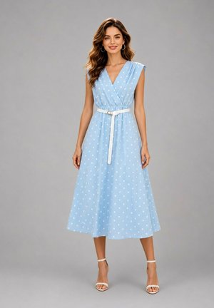 Femme portant une robe midi sans manches bleu clair à pois avec ceinture blanche et sandales blanches à talons hauts, debout devant un fond uni.