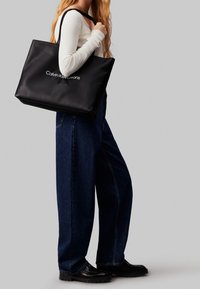 Czarna skórzana torba typu tote z białym logo "Calvin Klein Jeans", z mocnymi rączkami i przestronnym, prostokątnym kształtem.