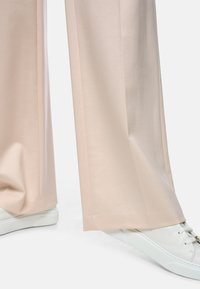 Close-up of beige wide-leg pants worn over white leather sneakers with laces and a small gold detail on the tongue.