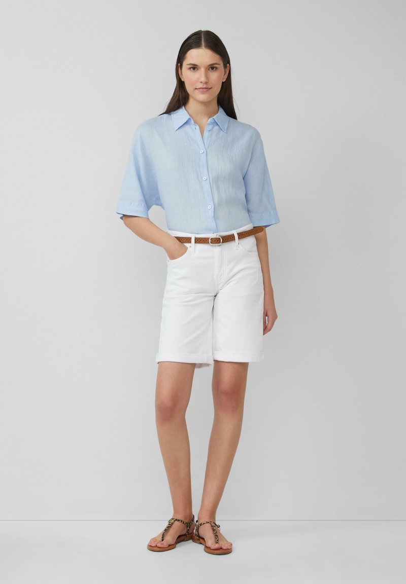 Helles blaues kurzärmliges Hemd mit einer weichen Textur, kombiniert mit weißen Shorts mit umgeschlagenem Saum und einem braunen geflochtenen Gürtel. Braune Sandalen.