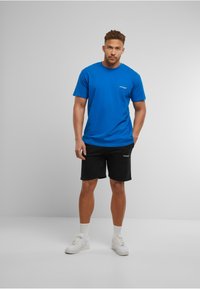 Blå bomulds t-shirt med rund hals og korte ærmer, kombineret med sorte shorts. Bemærkelsesværdig logo-detalje på brystet, hvide sneakers på fødderne.