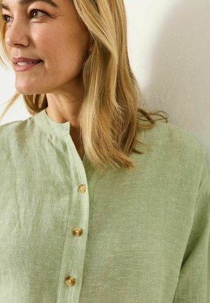 Sieviete ar plecu garuma blondiem matiem zaļganā linu kreklā ar trim redzamām pogām, skatoties uz kreiso pusi.