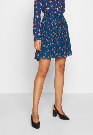 Femme portant une jupe plissée bleue fleurie et un top assorti à manches longues, associée à des chaussures slingback noires à talon bloc.