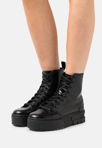 Botas altas negras hechas de material sintético brillante. Presentan suelas gruesas negras, cordones redondos y una marca en el lateral.