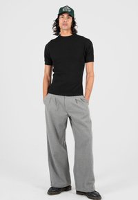 Uomo con tatuaggi che indossa una camicia nera a maniche corte, pantaloni grigi larghi, scarpe nere e un cappellino verde, in piedi con le mani nelle tasche.