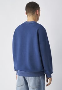 Felpa blu realizzata in tessuto morbido, con un design leggermente oversize, polsini a coste e collo tondo. Vista dal retro.