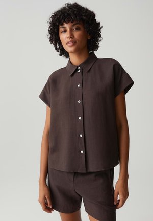 Frau mit lockigem Haar, die ein dunkelbraunes kurzärmeliges Hemd und passende Shorts trägt, steht vor einem schlichten hellen Hintergrund.