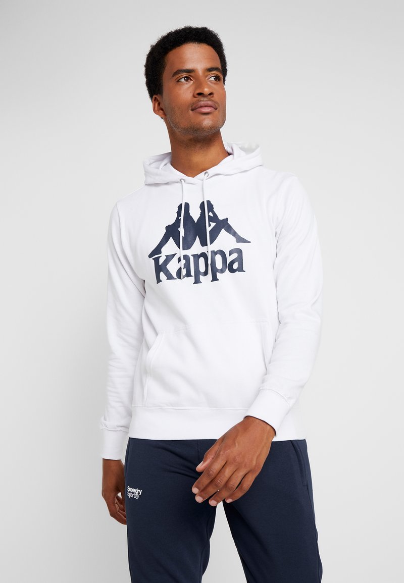 Hombre vestido con una sudadera blanca de Kappa con un logo negro y pantalones deportivos azul marino de Superdry Sport, mirando ligeramente de lado.