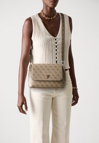 Borsa a tracolla beige con motivo, chiusura a patta, hardware logo dorato e tracolla texture. Indossata con un top senza maniche in maglia e pantaloni bianchi.