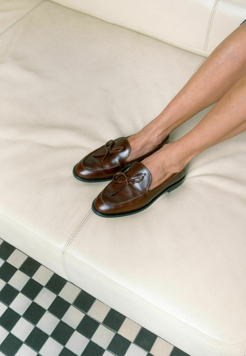 Bruine leren loafers met een ronde neus, met een strikaccent op de voorkant, geplaatst op een lichtgekleurde bank boven een vierkante vloer.