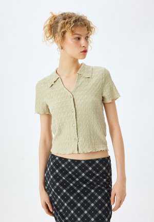 Camisa de botones de manga corta, verde claro, con un patrón texturizado y ondulado, dobladillo en forma de concha y cuello clásico. Combinada con una falda de cuadros.