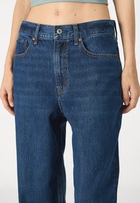 Pantalones vaqueros de mezclilla en azul oscuro con cintura alta, diseño de cinco bolsillos y costuras en contraste, que cuentan con un cierre de botón y cremallera.