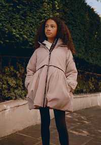 Aufgeblähte rosa Jacke mit tailliertem Bund und elastischen Bündchen. Verfügt über eine Kapuze und ein Logoband entlang des Reißverschlusses. Kombiniert mit schwarzen Leggings.