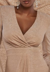 Kvinna iklädd en skimrande beige omwrap-klänning med rynk detaljer och hängande kristallkorsörhängen, mot en matchande glittrande bakgrund.