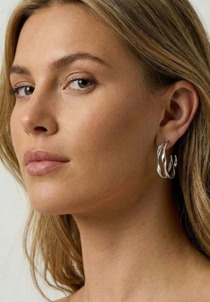 Woman with light brown hair wearing silver twisted hoop earrings, looking slightly to the side against a plain background.
