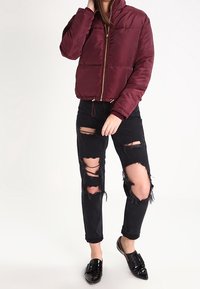Chaqueta de plumas burdeos con cuello alto y cremallera frontal, combinada con jeans negros desgastados con grandes desgarros y mocasines negros.