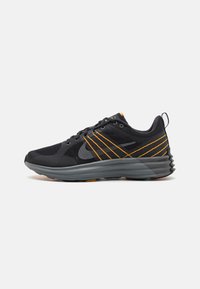 Des chaussures de sport noires avec une tige en mesh, présentant des accents orange et des détails texturés. La semelle est grise avec un design ondulé pour améliorer l'adhérence.