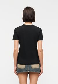 Γυναίκα με κοντά καστανά μαλλιά, που φοράει μια απλή μαύρη μπλούζα και μια κοντή τζιν φούστα με ταμπά τσέπες, γυρισμένη προς τα πίσω μπροστά σε λευκό φόντο.