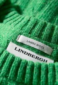 Un suéter de lana de cordero verde presenta una textura acanalada, con etiquetas que muestran "Lindbergh" y "Lana de Cordero" en blanco y negro.