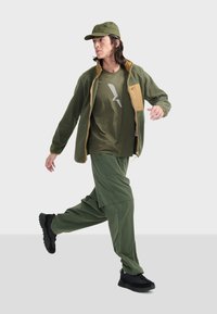 Grøn fleecejakke med beige detaljer, olivengrøn t-shirt med grafik, grønne bukser og sorte sneakers. Model i bevægelse.