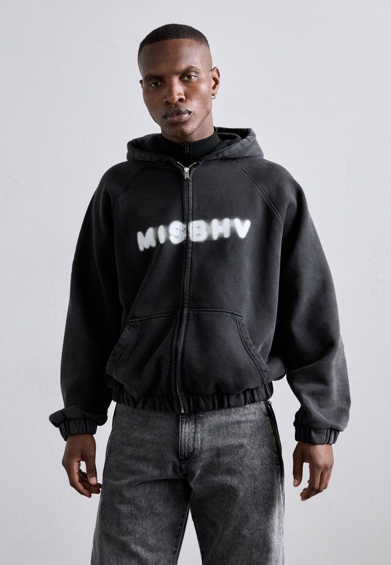 Schwarze Zip-Hoodie mit weißem "MISBHV"-Text. Verfügt über eine Kapuze mit Kordelzug, elastische Bündchen und Seitentaschen. Kombiniert mit grauen Jeans.