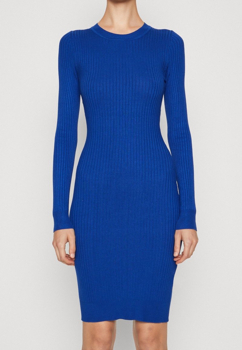 Femme portant une robe moulante en maille côtelée à manches longues, de couleur bleu cobalt vif, debout devant un fond clair uni.