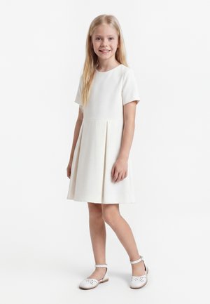 Ung pige smilende, iført en knælængde hvid kjole med korte ærmer og hvide ankelremssko, stående mod en ensfarvet baggrund.