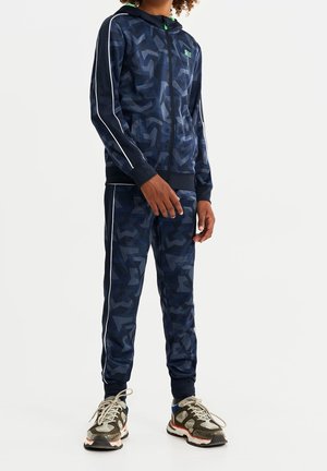 Tuta navy con motivo geometrico, strisce laterali bianche, felpa con zip e polsini a costine. Tessuto testurizzato, abbinato a sneaker chunky.