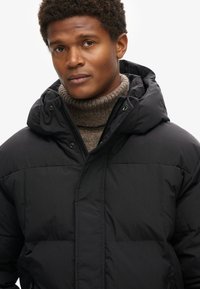 Giacca nera imbottita con cappuccio, dotata di zip frontale e chiusure a scatto, indossata sopra un maglione beige a collo alto. Texture liscia e design funzionale.