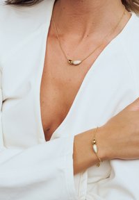 Guldkedja med två droppformade hängen, en vit med textur och en slät. Guldarmband med liknande hängdesign.