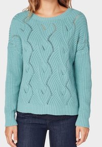 Pull bleu clair en tricot avec une texture côtelée, présentant des motifs ondulés et des détails en ajouré. Col rond et manches longues.