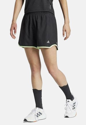 Pantalones cortos deportivos negros con ribete verde, cintura elástica y logo de Adidas. Combinados con zapatillas blancas y calcetas negras.