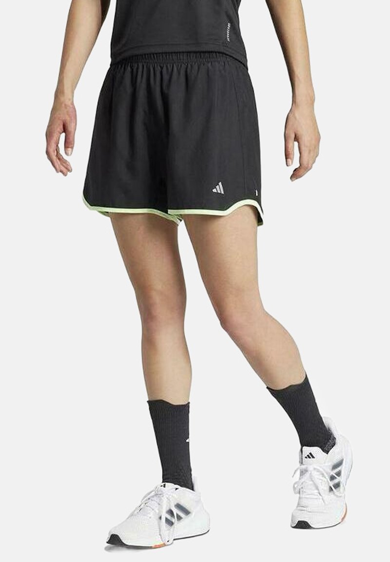 Pantalones cortos deportivos negros con ribete verde, cintura elástica y logo de Adidas. Combinados con zapatillas blancas y calcetas negras.