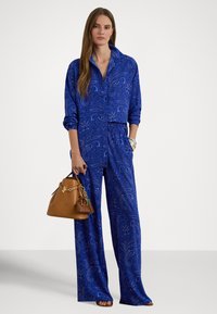 Γυναίκα που φορά μπλε μπλούζα με πασλέ μοτίβο και μακρυμάνικο σχέδιο, καθώς και παντελόνια με φαρδιά πόδια που ταιριάζουν, κρατώντας καφέ δερμάτινη τσάντα με χρυσές λεπτομέρειες.