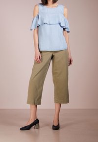 Kvinna i ljusblå kallaxel-top, olivgröna vida capribyxor och svarta klackskor som står mot en neutral bakgrund.