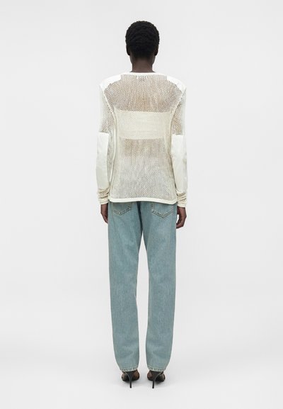 Personne à la peau foncée et aux cheveux courts bouclés, portant un haut blanc transparent à manches longues, un jean bleu clair et des chaussures noires à talons hauts, vue de dos.