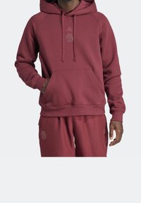 Homme portant un sweat à capuche Adidas bordeaux et un pantalon de survêtement assorti, la main dans la poche du sweat, debout devant un fond clair uni.