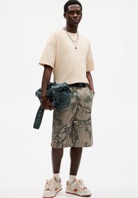 Beige oversize tílko, maskáčové šortky se zelenými akcenty, držící tmavě zelenou bundu a obuté bílé sneakers s oranžovými detaily.