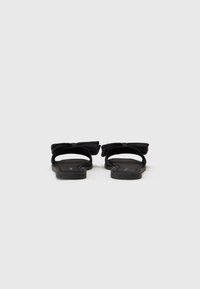 Sandale negre plate dintr-un material moale, cu un accent mare sub formă de fundă în spate. Design cu vârf descoperit, talpa plată oferă o elevare minimă.