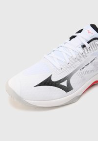 Chaussure de sport blanche avec des accents noirs et gris. Présente une tige texturée, des lacets plats et des détails rose pâle sur la semelle.